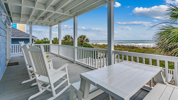 A covered outdoor deck with white railings and a table, overlooking a beach with people and palm trees. - Gorgeous Oceanfront Beach House, Outdoor Pool | Blue Heaven (North Myrtle Beach)