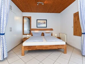 Un dormitorio con estructura de cama de madera, una mesita de noche con lámpara, un cuadro enmarcado en la pared y una ventana con cortinas.