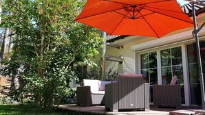 An outdoor patio with wicker furniture, a large orange umbrella, and a glass door leading to a covered area. - Holiday house Fichtenwalde, 160m², 2.5 BZ, 3SZ, WZ, dining room, kitchen, garden, VDSL (Beelitz)