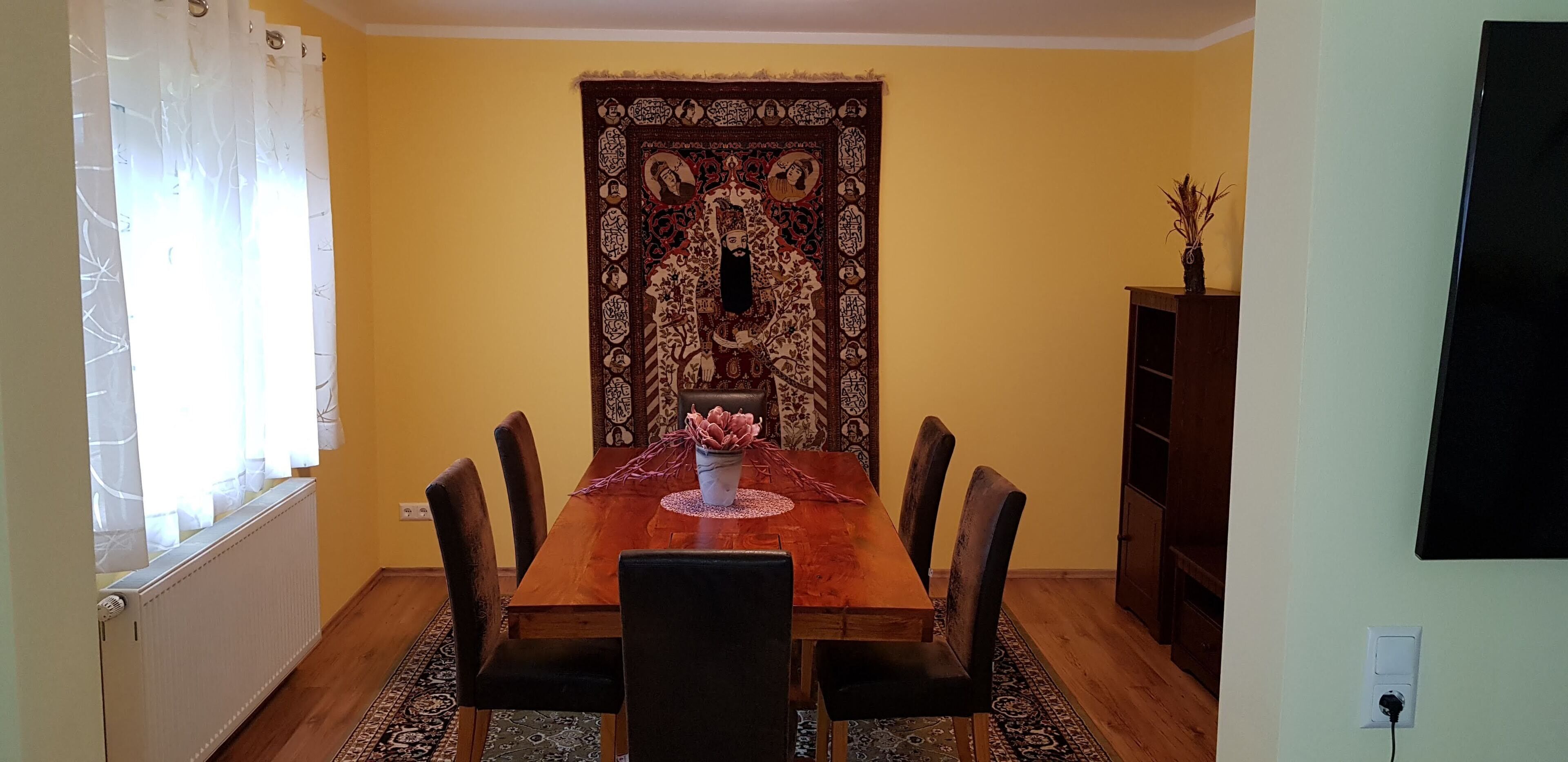 Une salle à manger avec une table en bois, six chaises, un vase de fleurs et une tapisserie au mur.