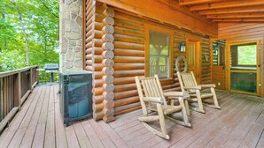 Una baita di legno con un portico, due sedie a dondolo, un muro di pietra e un barbecue.