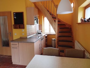 A compact kitchen with a wooden staircase, a dining area with a table, and a window with blinds.