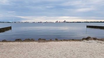 Ein Sandstrand mit ruhigem Wasser, eine ferne Stadtansicht und eine Brücke im Hintergrund.