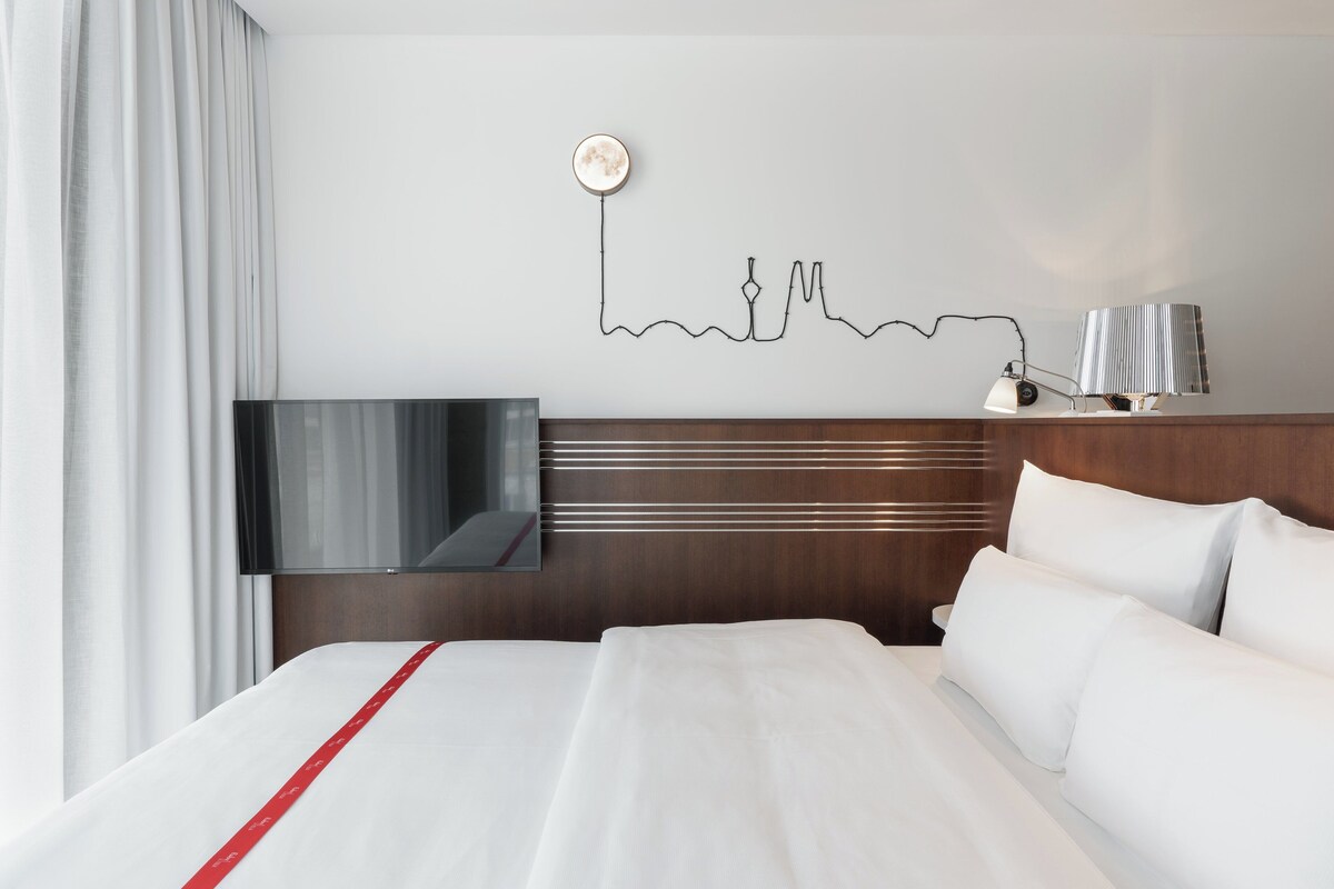 Habitación de hotel con cama, televisión montada en la pared, una obra de arte en la pared y mesita de noche con lámpara.