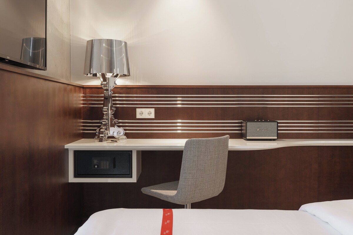 A hotel room with a bed, a desk with a lamp, a chair, and a small speaker.