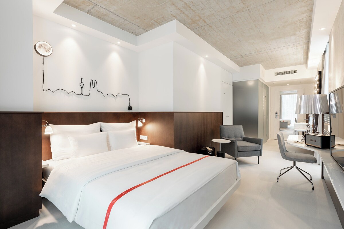 A modern hotel room with a large bed, a desk, a chair, and a wall art piece.