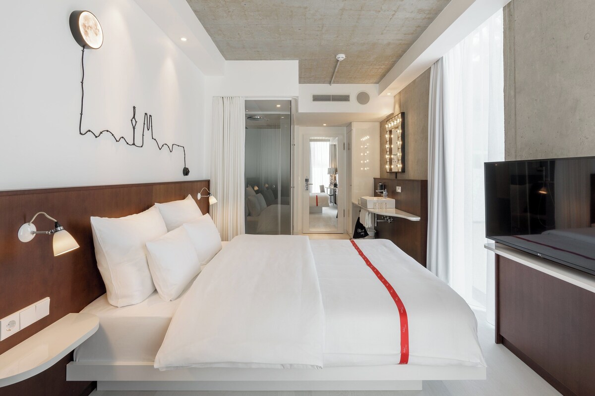 A modern hotel room with a large bed, a TV, a bathroom with a mirror, and a city skyline artwork on the wall.