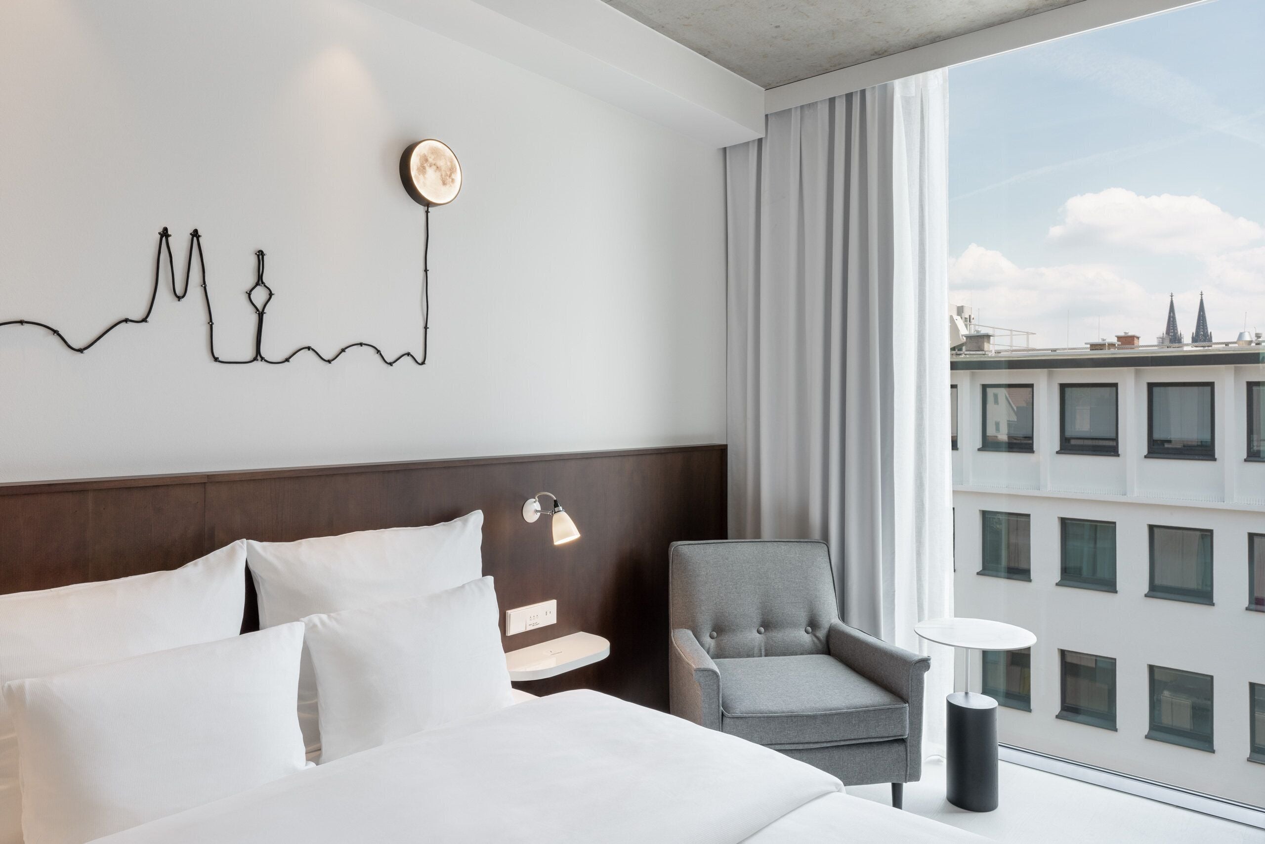 A hotel room with a bed, a chair, a small table, a wall art piece, and a window with a city view.