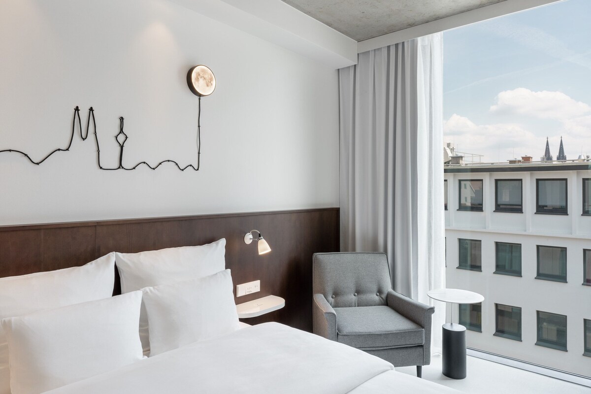 A hotel room with a bed, a chair, a small table, a wall art piece, and a window with a city view.