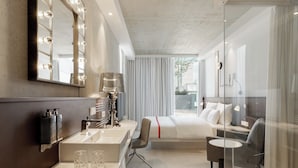 A modern hotel room with a large bed, a desk with a lamp, a vanity with a mirror and lights, and a view of the outdoors. - Ruby Ella Hotel Cologne by IHG (Cologne)