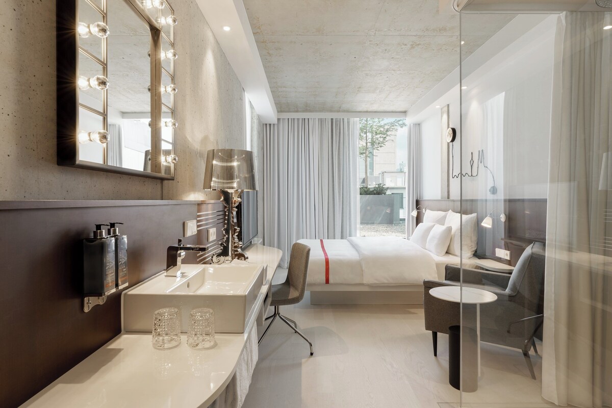 A modern hotel room with a large bed, a desk with a lamp, a vanity with a mirror and lights, and a view of the outdoors.