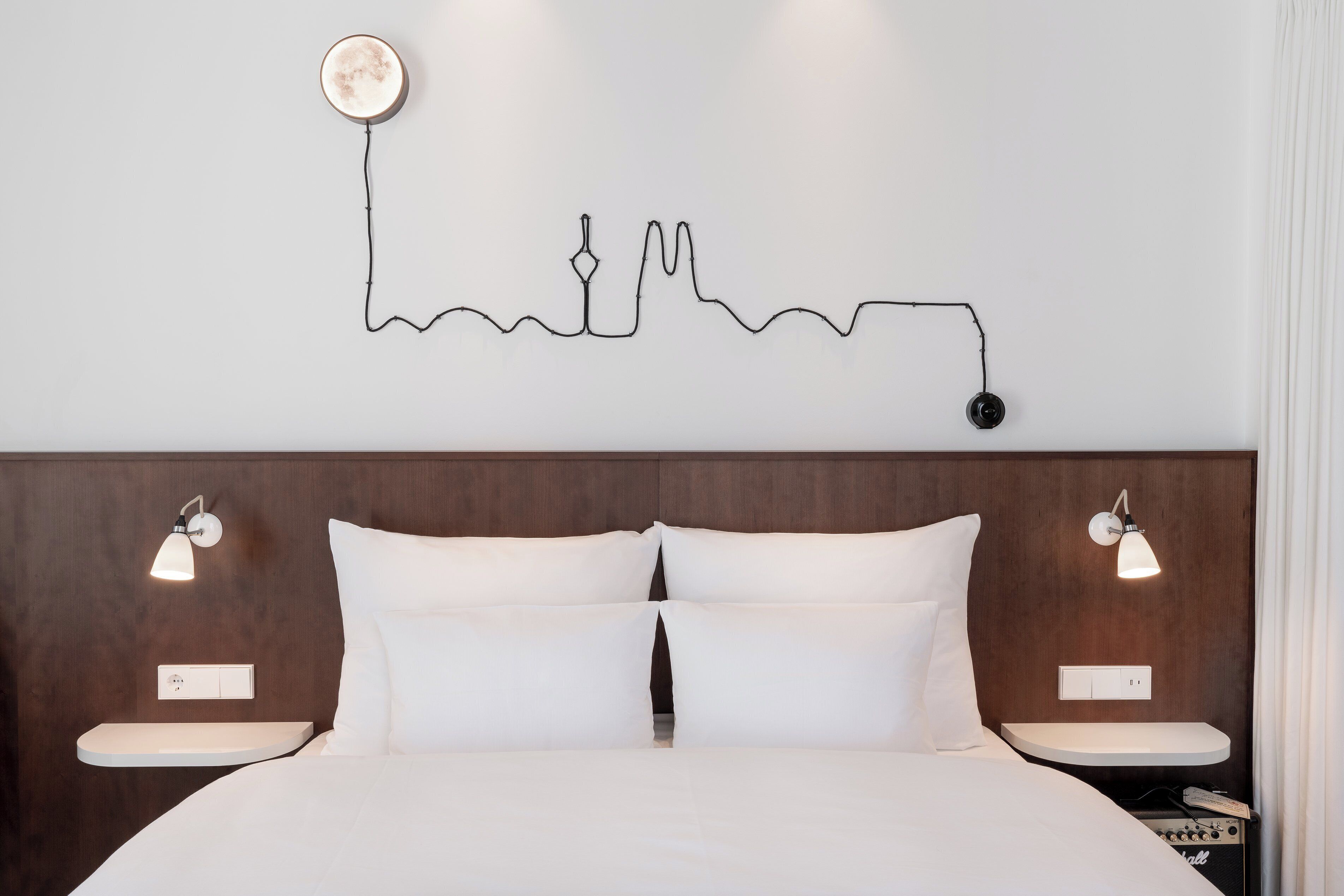 A hotel room with a bed, two bedside tables, a wall-mounted light fixture, and a wall art piece.