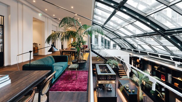 A modern hotel interior with a high ceiling, a dining area, a seating area with a sofa, and a staircase leading to an upper level. - Ruby Ella Hotel Cologne by IHG (Cologne)