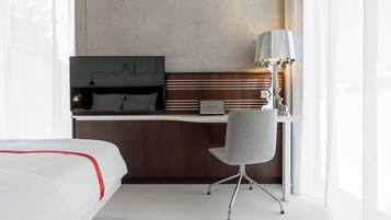 Quarto de hotel com uma cama, escrivaninha, cadeira, luminária e televisão presa na parede.