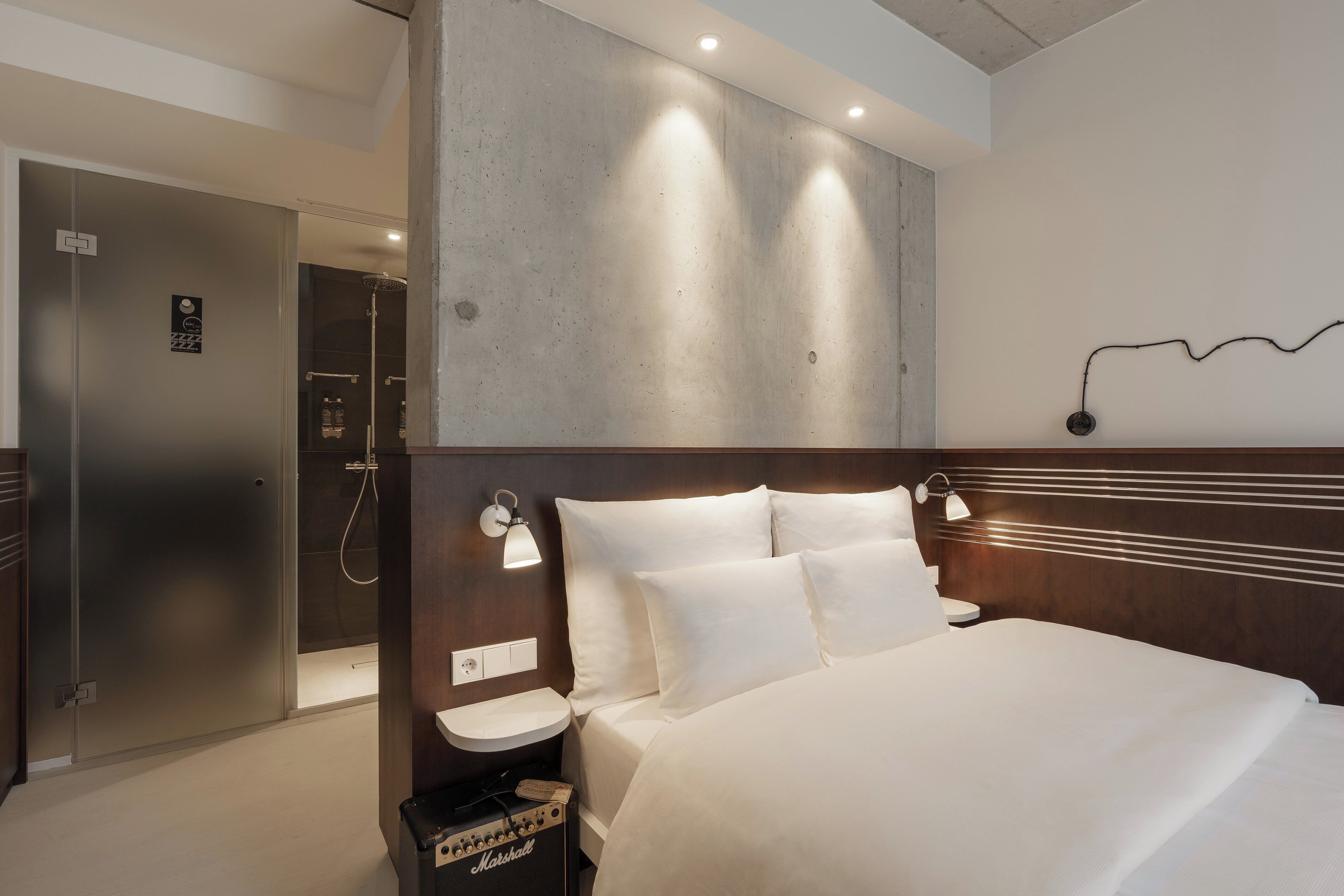 A hotel room with a bed, bedside table, lamp, and a shower area.