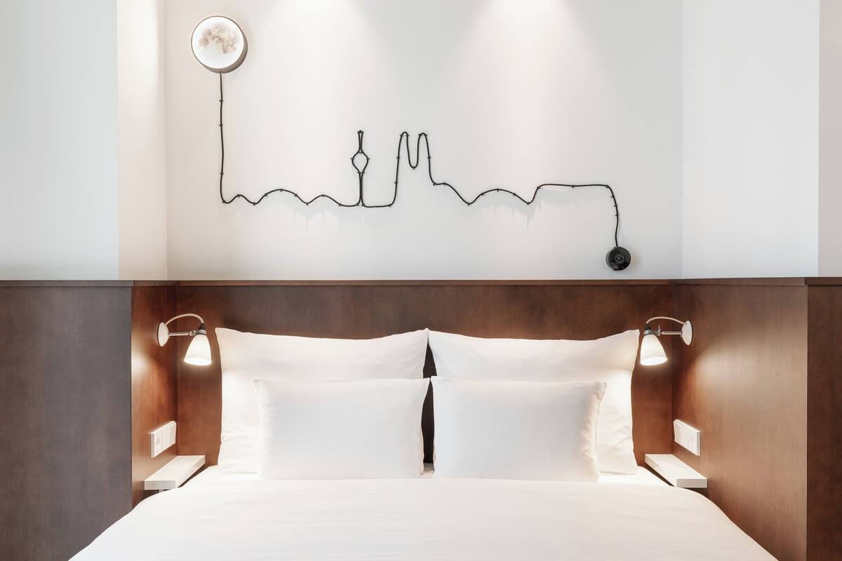 Quarto de hotel com cama, abajures, luminária na parede e um desenho simples adornando a parede.