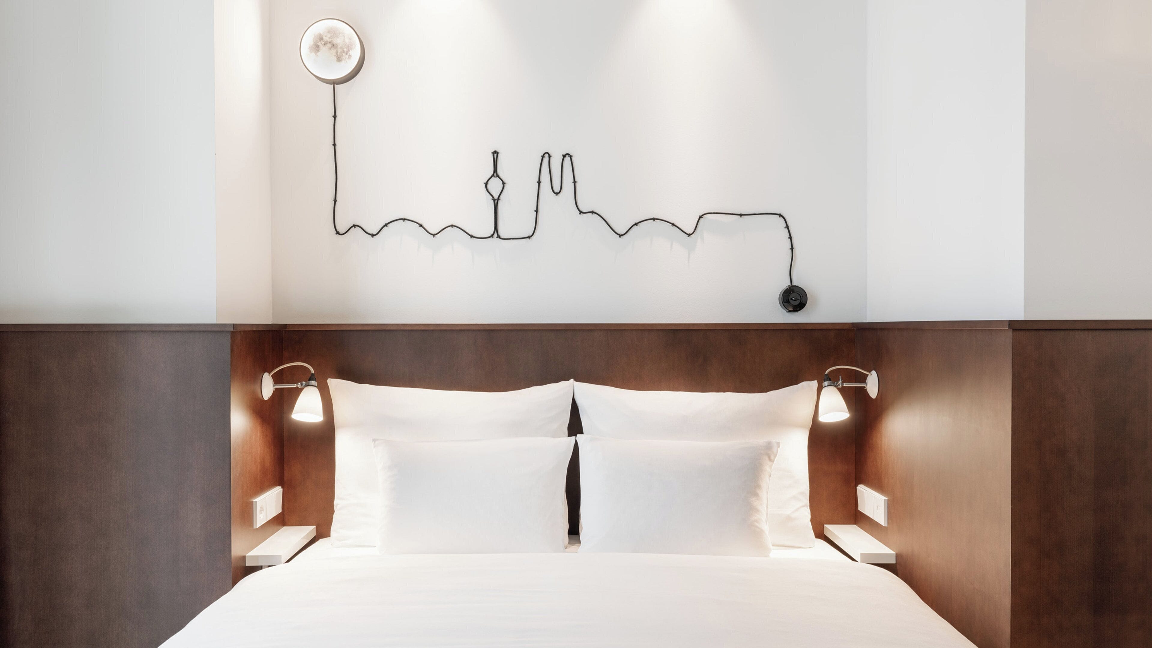 A hotel room with a bed, bedside lamps, a wall-mounted light fixture, and a simple line drawing on the wall.