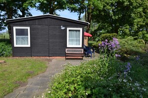 Une petite cabane noire avec deux fenêtres, un banc et une chaise à l’extérieur.