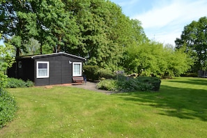 Une petite cabane à un seul étage, avec un banc à l’extérieur, entourée d’une végétation luxuriante et d’arbres.