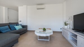 Ein modernes Wohnzimmer mit grauem Sofa, rundem Glastisch, Flachbildfernseher und einer weißen Klimaanlage an der Wand.