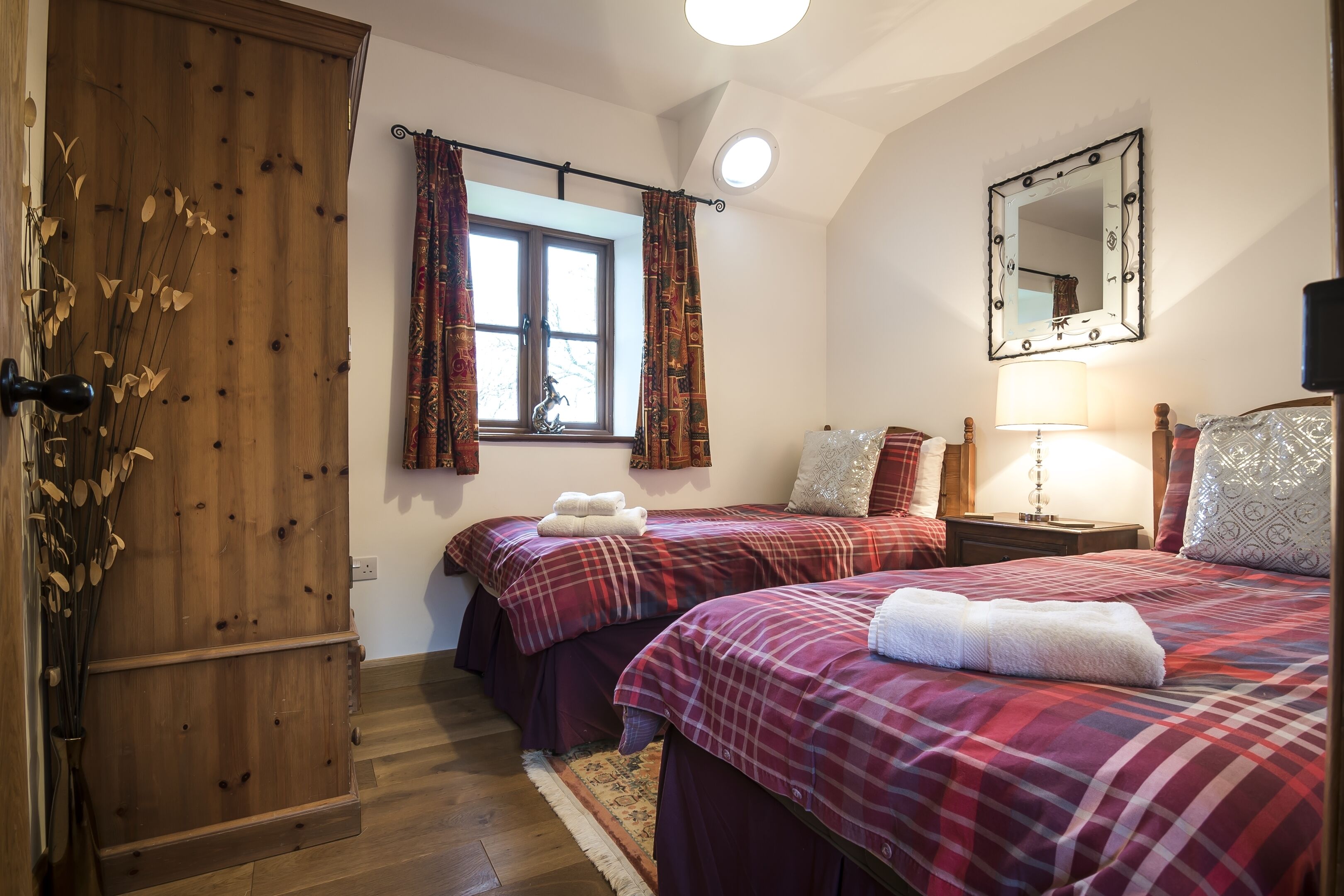 A bedroom with two beds, a wooden wardrobe, a window with curtains, a mirror, and a bedside lamp.