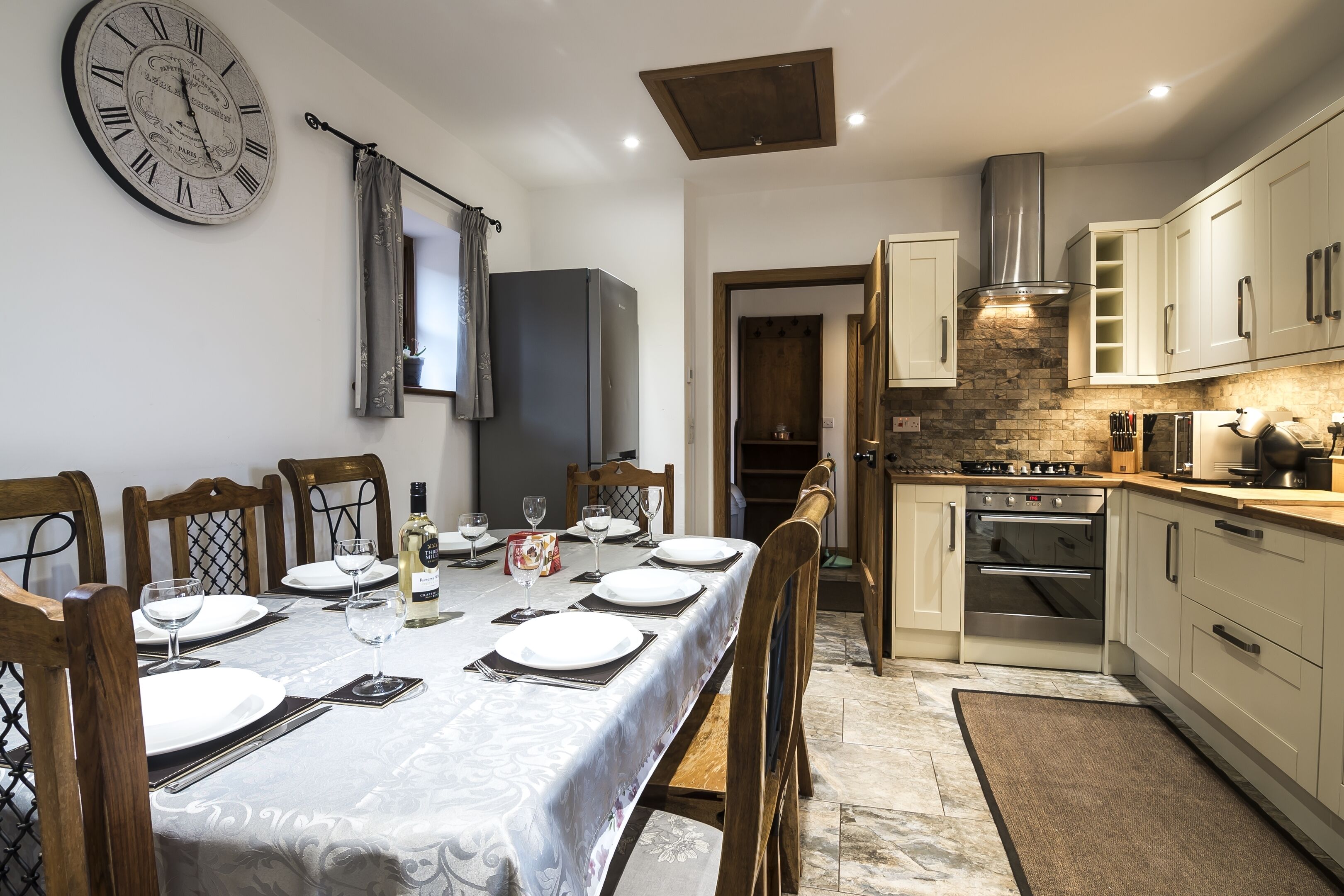 A dining area with a table set for four, a clock on the wall, and a kitchen with modern appliances.