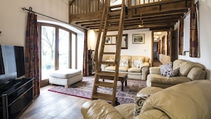 A spacious living room with a wooden staircase, leather sofas, a flat-screen TV, and a large window with curtains. - Orchard Cottage - Luxury Barn Conversion (Tenby)