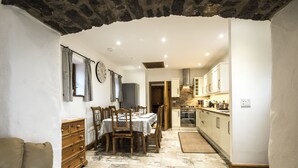 A dining area with a stone floor, wooden furniture, and a kitchen in the background. - Orchard Cottage - Luxury Barn Conversion (Tenby)
