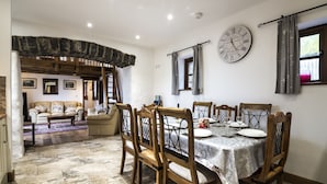 A dining area with a wooden table set for four, surrounded by wooden chairs. A large wall clock and a window with curtains are visible. - Orchard Cottage - Luxury Barn Conversion (Tenby)