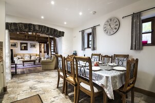 Un comedor con una mesa de madera dispuesta para cuatro personas, rodeada de sillas de madera. Se observa un reloj de pared grande y una ventana con cortinas.