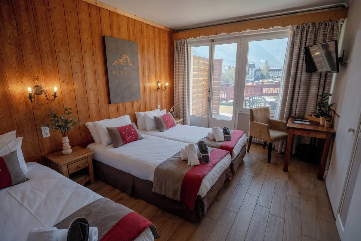 Habitación de hotel con paredes de madera, dos camas, un escritorio y un ventanal con vistas a montañas.