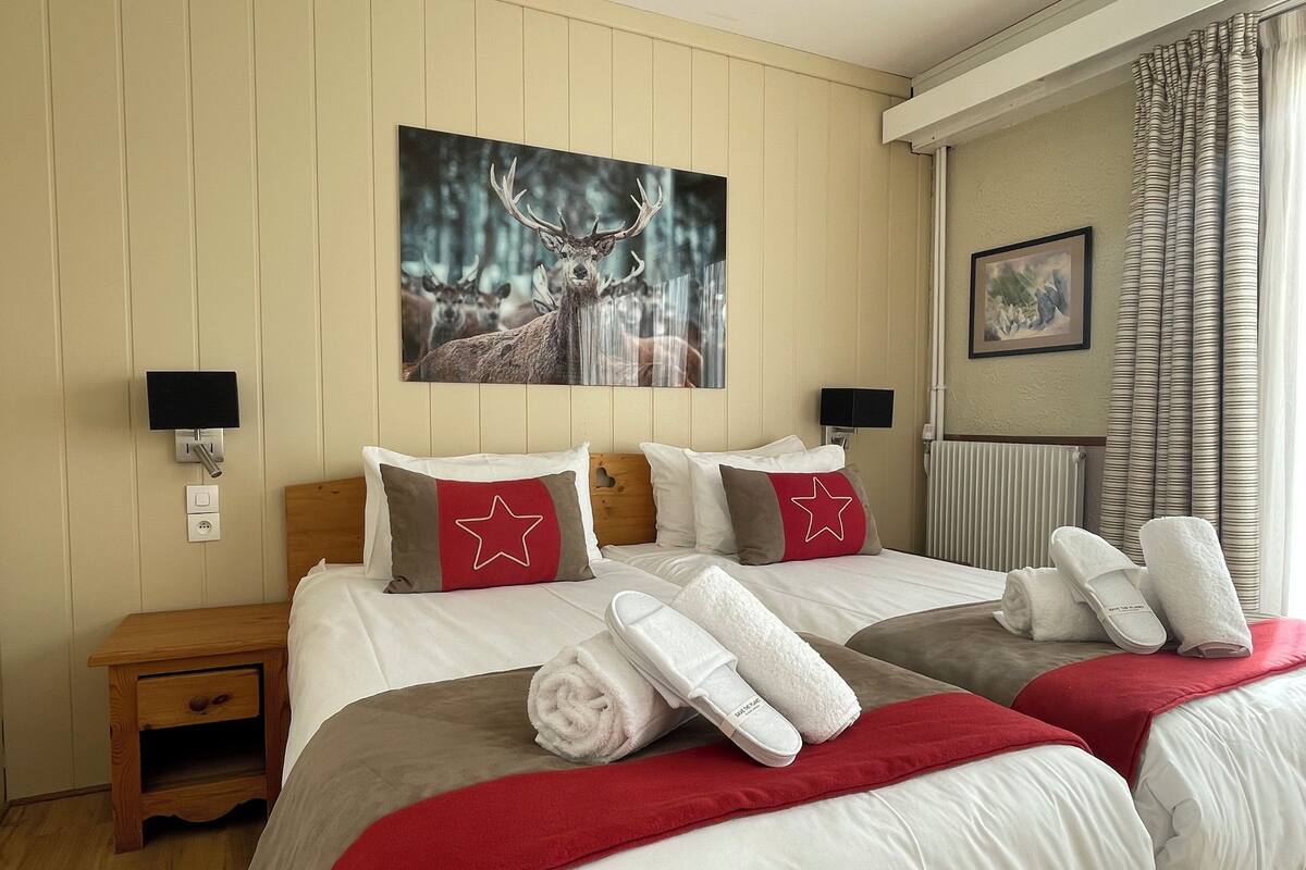 Habitación de hotel con dos camas, un cuadro de ciervos, un cojín con forma de estrella roja y una manta roja.