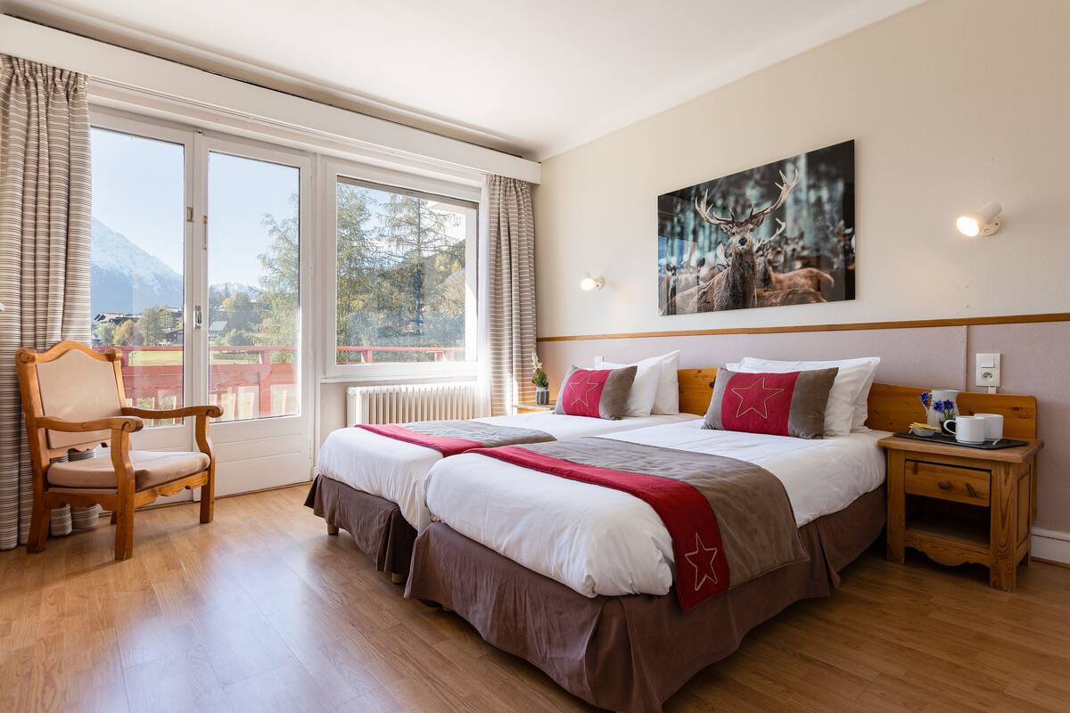 Habitación de hotel con una cama grande, una silla de madera, una mesita de noche y una ventana con vistas a las montañas.