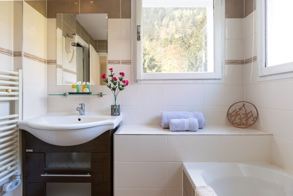 Un baño con lavamanos blanco, espejo y ventana. Hay un perchero con toallas y un adorno en la pared.