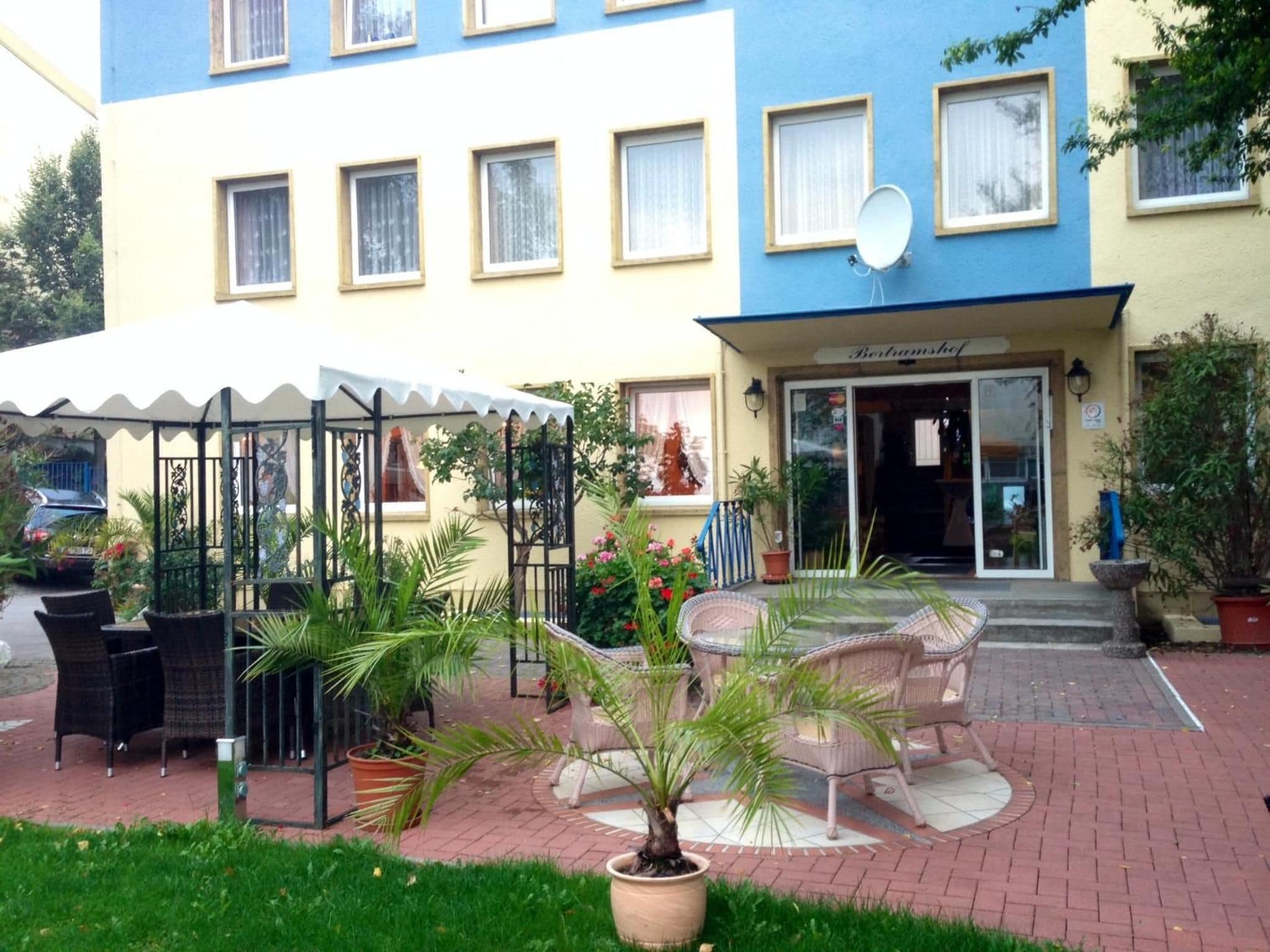 Ein Hotel Eingangsbereich mit überdachter Außenbestuhlung, Blumentöpfen und freiem Blick auf die Eingangstür.