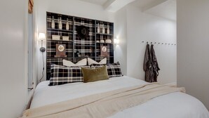Un dormitorio con cabecera a cuadros, cama con ropa de cama blanca, manta color beige y una lámpara fijada a la pared.