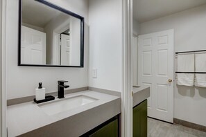 Un baño con lavamanos blanco, grifo negro y un espejo. Hay un perchero con toallas blancas en la pared.