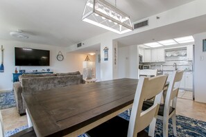 A modern living room with a large wooden dining table, a flat-screen TV, a sofa, and a kitchen in the background. - 🦩Book Me Baby! Endless Summer at The Enclave 204B~Heart of Destin Location🦩 (Destin)