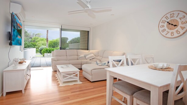 A modern living room with a sofa, a dining table, and chairs. There is a TV mounted on the wall and a ceiling fan. - Beachfront on Bennetts | Beaches Apartment 4 (Hawks Nest)