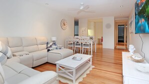 A modern living room with a beige sofa, a white coffee table, and a dining area with white chairs and a table. - Beachfront on Bennetts | Beaches Apartment 4 (Hawks Nest)