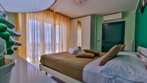 Een netjes opgemaakt bed met een bruine deken, witte kussens en een klein handdoekje. Een raam met dunne gordijnen laat natuurlijk licht de kamer binnen.