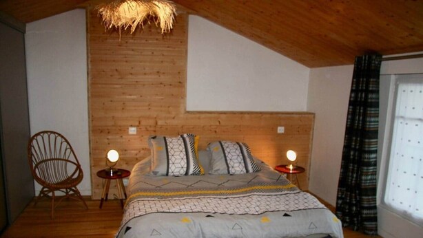 Une chambre avec un plafond en pente, un lit avec une literie à motifs, deux tables de chevet avec des lampes, un fauteuil en osier et une fenêtre avec des rideaux.