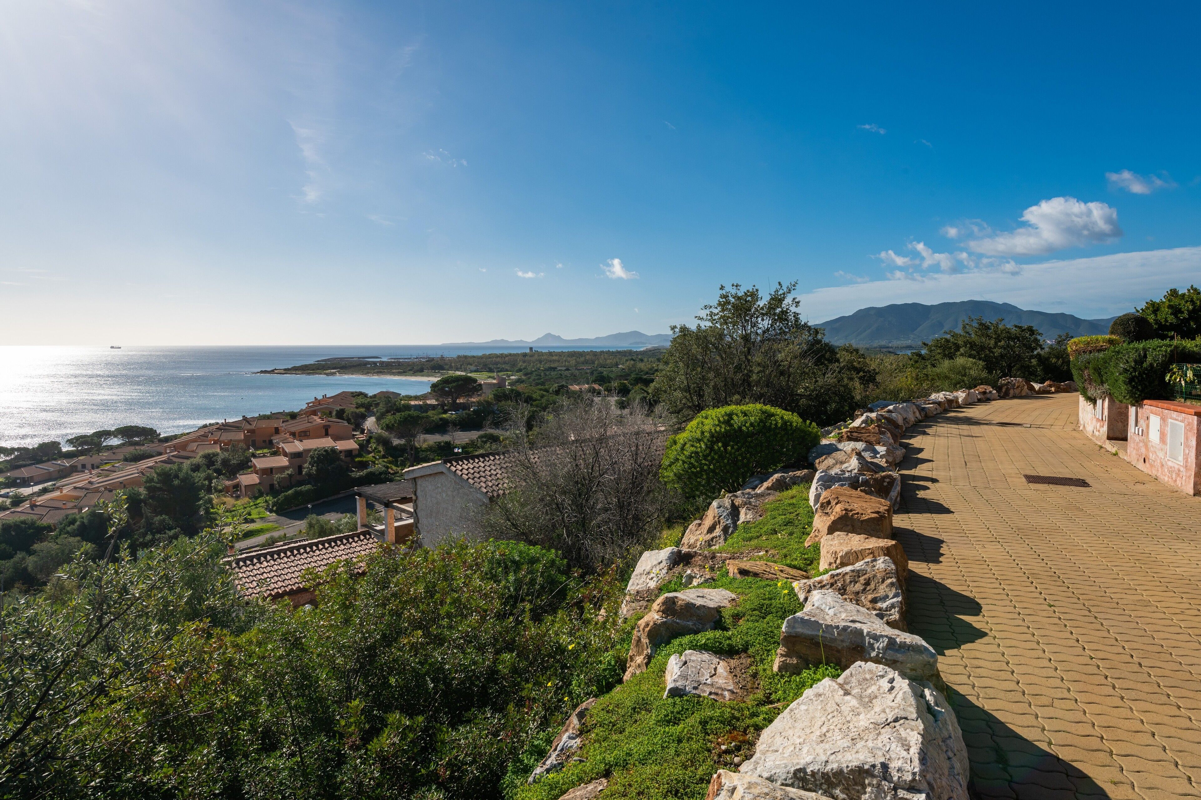 Una località costiera con edifici dalle tegole color terracotta, un viale lastricato e vista sul mare e sulle montagne.