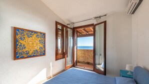 A bedroom with a bed, a nightstand, a lamp, a large window with a view of the sea, and a wall-mounted air conditioner.
