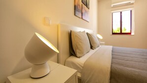 Une chambre à coucher bien rangée, avec un lit, une lampe de chevet, une table de nuit et une fenêtre laissant entrer la lumière du jour.