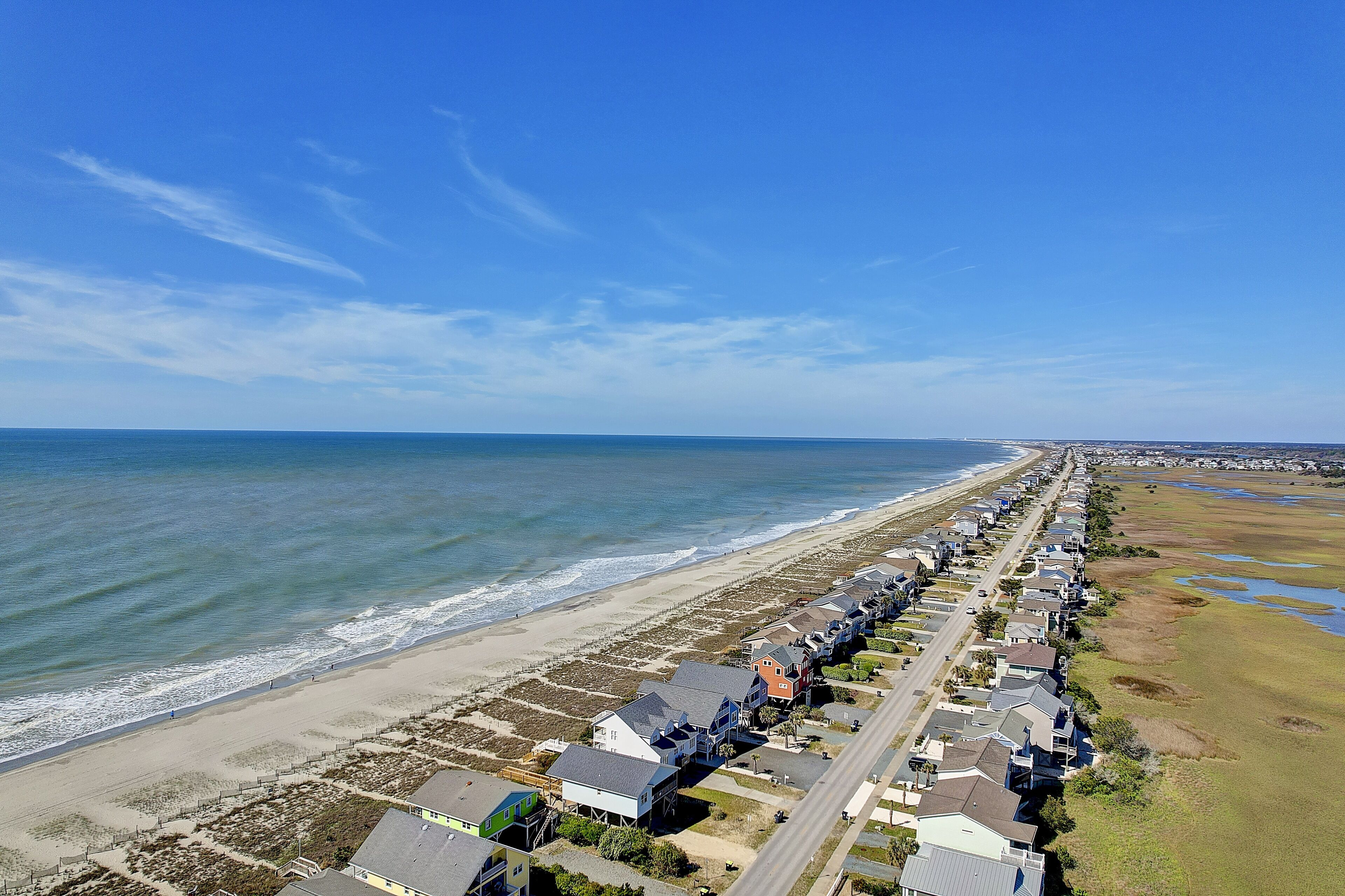 A coastal residential area with houses along the beach, a clear sky, and a road running parallel to the shore.