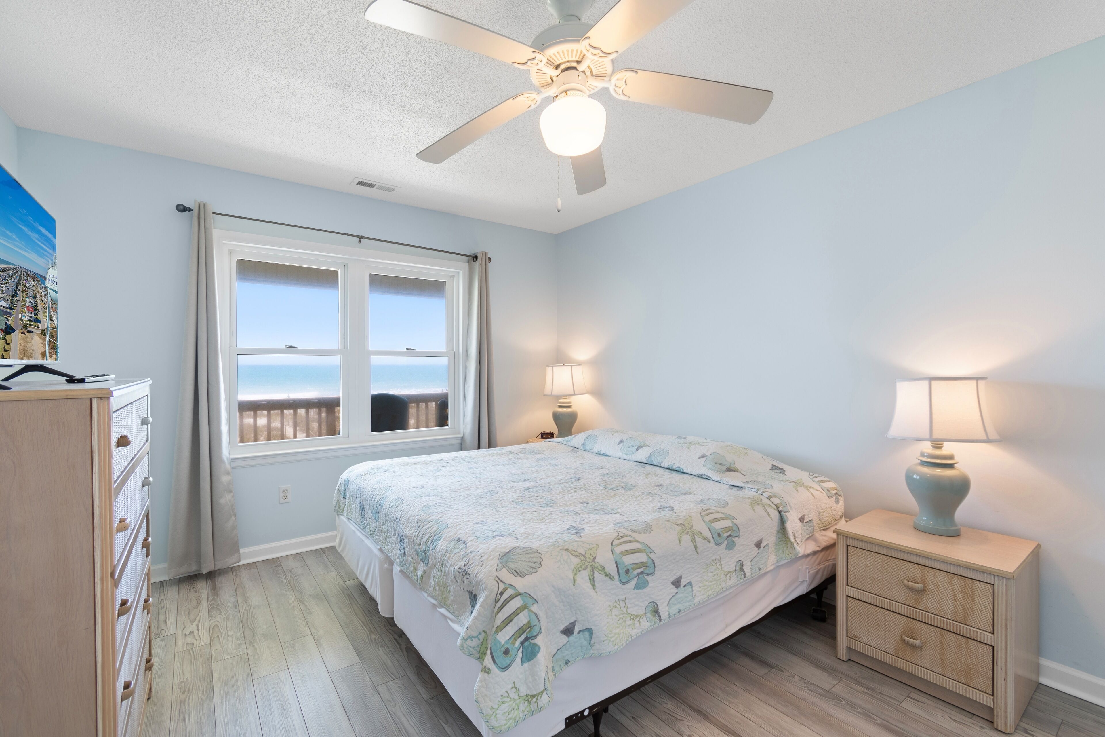 A bedroom with a bed, a nightstand, a dresser, a ceiling fan, and a window with a view.