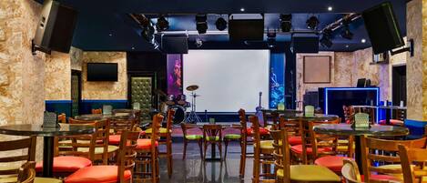 A restaurant with multiple tables and chairs, a stage with musical instruments, and a large screen.