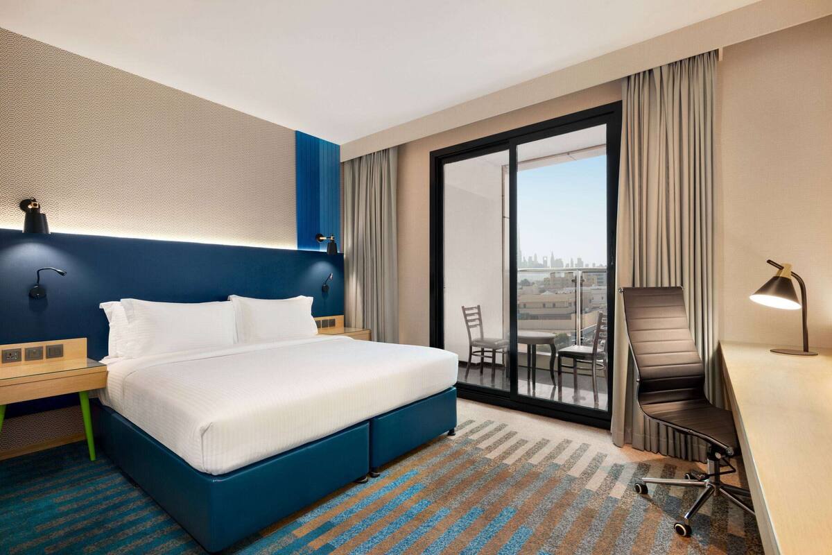 Habitación de hotel con una cama grande, un escritorio con una silla, una computadora portátil, una lámpara de escritorio y vistas de la ciudad a través de una puerta corrediza de vidrio.