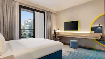 A hotel room with a large bed, a desk with a computer, a TV, and a view of buildings through the window.